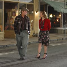 a man and woman walking down the street at night in front of a storefront