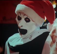 a man in a clown mask and santa hat with his face painted white, standing next to a teddy bear