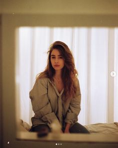 a woman sitting on top of a bed in front of a window with white curtains