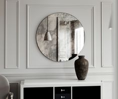 a round mirror hangs on the wall above a white cabinet with drawers and a vase