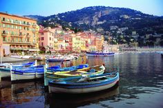 there are many small boats docked in the water near some buildings and hills behind them