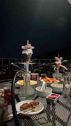 an outdoor table with food and drinks on it, overlooking the city lights at night