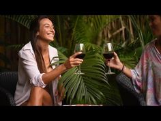 two women sitting at a table with wine glasses in their hands and palm trees behind them