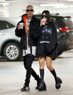 a man and woman walking through an airport holding coffees in their hands, both wearing black over - the - knee boots