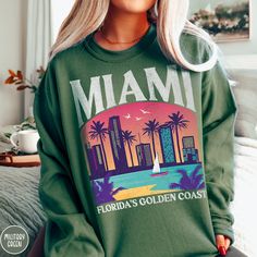 "Celebrate \"Vice City\", the capital of South Florida, the gem of Florida's Golden Coast, the Sunshine City.... MIAMI FLORIDA, with our one of kind Triangle Threads Shop Miami Sweatshirt. This crewneck is perfect for those cold nights on the beach, or warming up after a dip in the pool back at the hotel. Celebrate your bachelorette, your family trip, or a Florida family reunion with this unisex sweatshirt perfect for all styles and ages. The Gildan 18000 50/50 blend fabric has that comfy, lived-in feel, that's like a hug every time you put it on! Now you can take a bit of Miami with you wherever you go! Grab one today! * Product Highlights * Ideal for any situation, this Gildan 18000 heavy blend crewneck sweatshirt is pure comfort. Made from an extremely soft 50/50 polyester and cotton bl Miami Beach Tshirt, Bebesota Sweater, Florida Sweatshirt, Beach Crewneck, Sunshine City, Miami Bachelorette, Miami Vacation, Golden Coast, Beach Sweatshirt