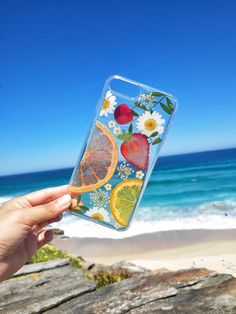 a person holding up a phone case with fruit and flowers on it, in front of the ocean