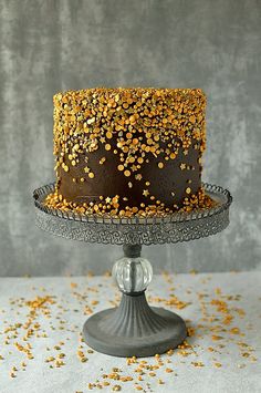 a chocolate cake with gold sprinkles on a metal stand against a gray background