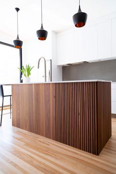 an island in the middle of a kitchen with wooden flooring and hanging lights above it