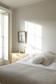 a bed with white sheets and pillows in a bedroom next to two windows that have light coming through them