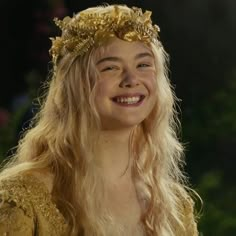 a woman with long blonde hair wearing a gold crown and smiling at the camera while standing in front of flowers