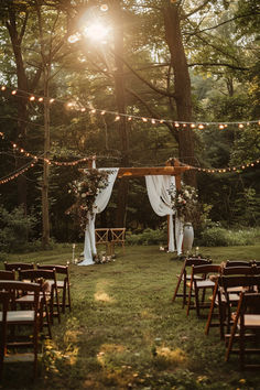 A beautiful garden wedding setup with a wooden arch draped in white fabric and adorned with floral arrangements, surrounded by string lights and wooden chairs under tall trees. Forest Backyard Wedding, Wedding In The Garden Decoration, Outdoor Wedding Lighting Ideas Backyards, Backyard Ceremony Decor, Wedding By Pond Ideas, November Wedding Outdoor, Backyard Budget Wedding, Dreamy Outdoor Wedding, Small Ceremony Big Reception