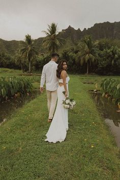 a bride and groom walking in the grass near some water with mountains in the background