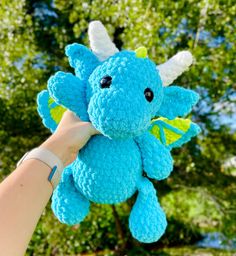 a hand holding a blue crocheted dragon stuffed animal in front of some trees