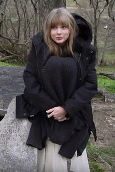 a woman standing next to a large rock in the woods, wearing a black coat and white pants