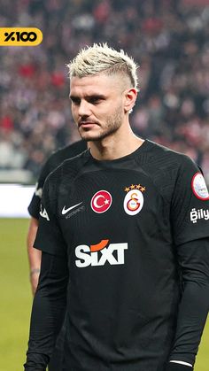 a man with blonde hair standing on a soccer field wearing a black shirt and shorts