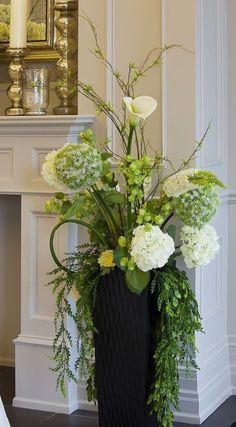 a black vase with white flowers and greenery in front of a fireplace mantel