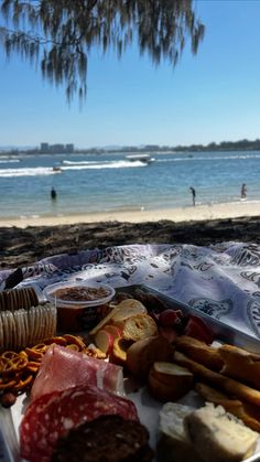 an assortment of cheeses, meats and bread on a picnic blanket at the beach