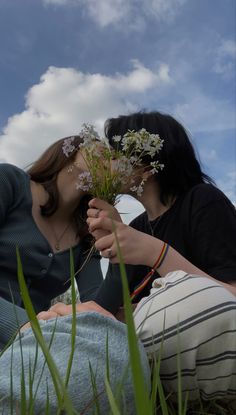 two women sitting on the grass kissing each other with flowers in their hands and blue sky behind them