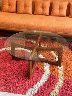 a glass table sitting on top of a rug in front of a couch with orange pillows