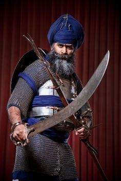 a bearded man dressed in medieval clothing holding two large swords and wearing a blue turban