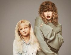 two women standing next to each other in front of a gray background, one with long blonde hair