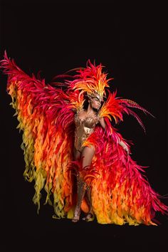 a woman in a colorful costume with feathers on her head and arms, standing next to a black background