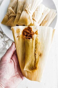 a person holding up a tamales on a white plate next to other food items