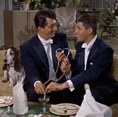 two men in suits sitting at a table with food on it and a dog standing next to them