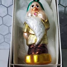 a christmas ornament in a box with a gold base and green hat on top