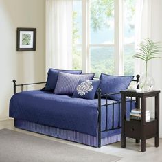 a day bed with blue sheets and pillows in front of a window, next to a night stand