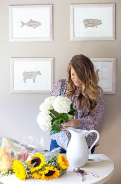 a woman arranging flowers on a table in front of three framed pictures and a vase