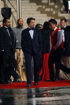 men in tuxedos stand on a red carpet as people watch from the steps