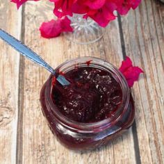 a jar filled with jam next to red flowers on a wooden table and a knife sticking out of it