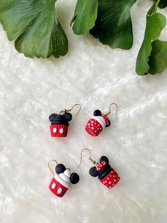 three red and white mickey mouse earrings on top of a white furnishing next to green leaves