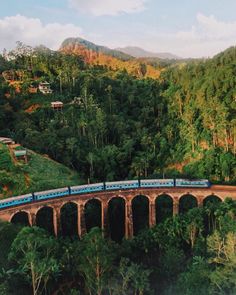 a blue train traveling over a bridge surrounded by lush green trees and mountains in the background