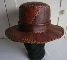 "Hand crafted leather brimmed hat in patchwork of wine coloured tones, with feature hand stitching in brown waxed thread.  Rim circumference approximately 53 cm / 21″. Height of crown is about 11 cm / 4.5″. Brim is about 8 cm / 3\" deep.  Please measure to check the hat circumference and height size you need before ordering, or contact me with any queries. Wine coloured leather band about 4 cm /1.5\" high around base of crown, with small circular motif and brass stud on one side.  Band is detachable if preferred. The hat is constructed in off-cut/up-cycle leather. The crown is lined in black fabric and brim is lined in black canvas. Handmade leather label inside crown \"Handmade by Reynold Trim\", who is the Entertainer Trim Bling Star." Brown Leather Fedora Hat, Fitted Brown Cap, Brown Hats With Leather Lining And Curved Brim, Brown Curved Brim Hat With Leather Lining, Distressed Brown Leather Brimmed Hat, Vintage Distressed Brown Leather Hat, Distressed Brown Leather Fedora Hat, Distressed Brown Leather Fedora, Brown Leather Hats For Festivals