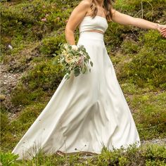 a woman in a white wedding dress holding her hand out to the side while standing on a hill