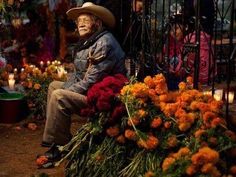 an old man sitting on a bench with flowers and candles in front of him at night