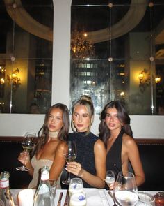 three beautiful women sitting at a table with wine glasses in front of them, posing for the camera