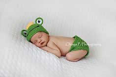 a baby wearing a crocheted frog hat and diaper is sleeping on a bed