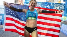 a woman is holding an american flag and posing for the camera while standing in front of a marathon finish line