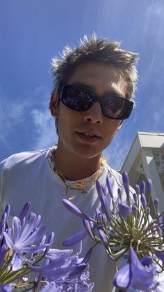 a man wearing sunglasses standing next to purple flowers in front of a blue sky with white clouds