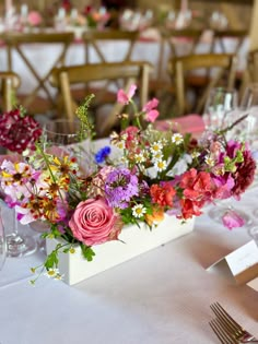 A low ceramic trough filled with abundant naturally styled multicoloured flowers sits on a table with a white table cloth set up for a wedding breakfast with cutlery and glasses. Colourful Flowers Table Setting, Colorful Wedding Table Centerpieces, Colourful Centerpieces Wedding, Colourful Table Centre Pieces, Simple Colourful Wedding Decor, Bright Wedding Flowers Centerpieces Table Decorations, Bright Floral Arrangements Wedding, Wedding Table Centerpieces Colourful, Bright Flowers Wedding Table