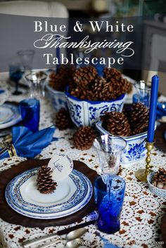 a table set with blue and white dishes, pine cones, silverware and candlesticks