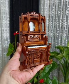 a hand holding a miniature wooden piano in front of a window with curtains behind it