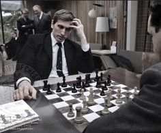 a man in a suit playing chess with another man sitting at a table behind him