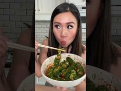 a woman holding a bowl filled with green peppers and chopsticks in her mouth