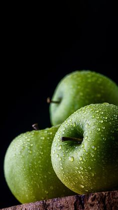 two green apples sitting on top of a piece of wood with water droplets covering them