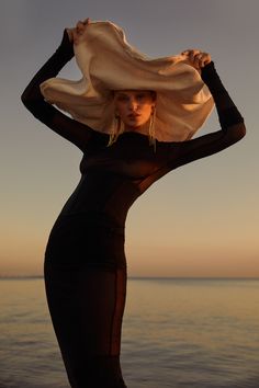 a woman in a black dress and hat standing by the water with her hands on her head