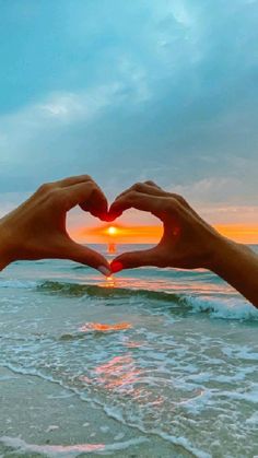 two hands making a heart shape with the sun setting in the background over the ocean